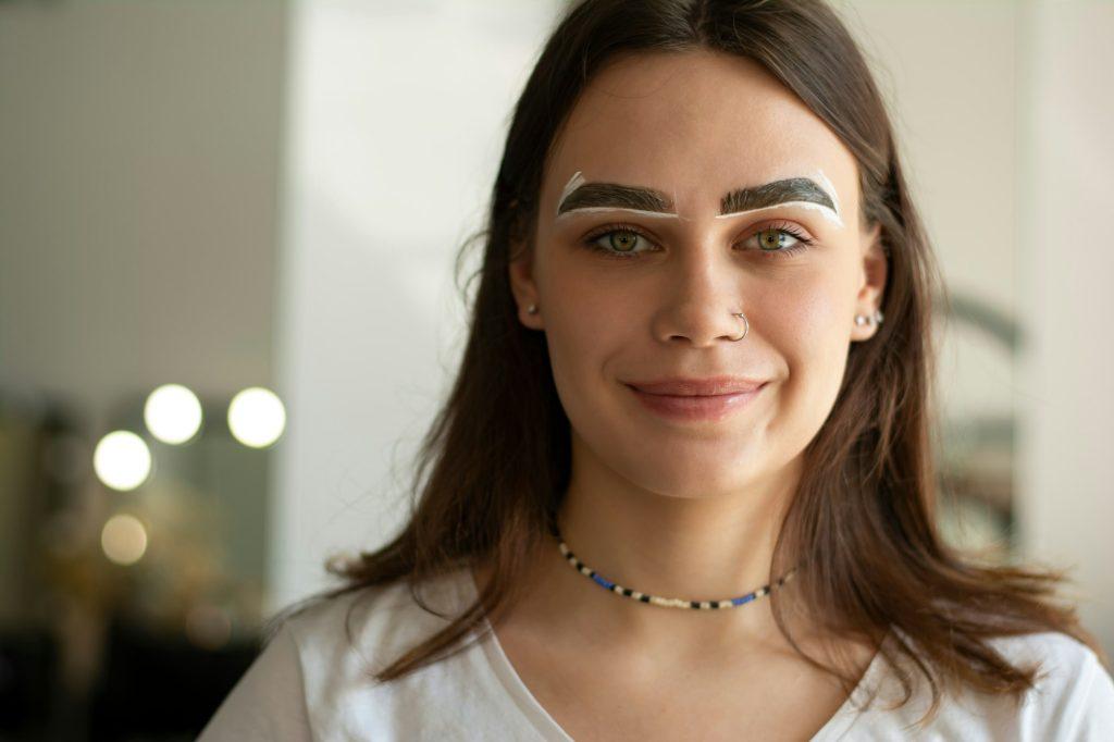 The process of dyeing the eyebrows of a young brunette who is looking directly at the camera